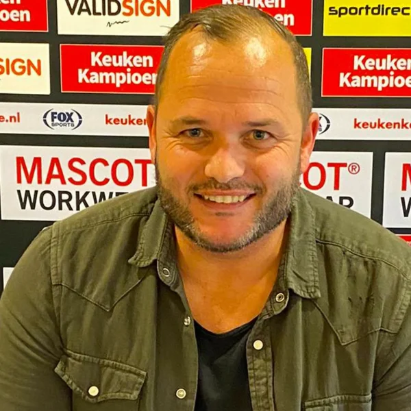 Nieuwe trainer Rico Goffin van FC Gulpen poseert lachend voor een sponsorwall in het Roda JC stadion
